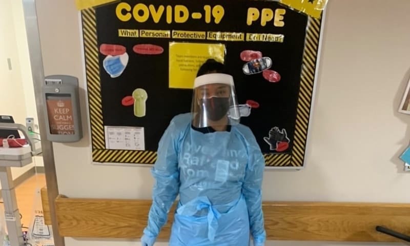 A nurse in blue scrubs and full personal protective equipment stands in front of a bulletin brad that reads ‘COVID-19 PPE.’