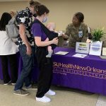 Two smiling women wearing military fatigues stand behind a table that displays a sign-up sheet, shirt and signs for the SVA. The table is draped with a deep purple cloth that says LSU Health New Orleans Health Sciences Center School of Nursing. One of the women is speaking to a young man and woman on the other side of the table, the latter of whom is holding an infant.