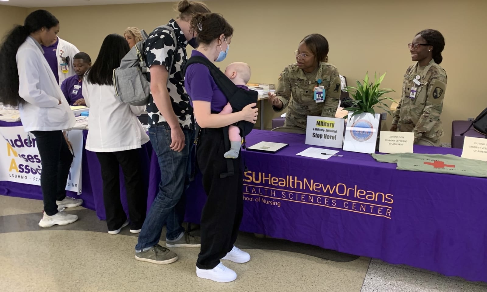 Two smiling women wearing military fatigues stand behind a table that displays a sign-up sheet, shirt and signs for the SVA. The table is draped with a deep purple cloth that says LSU Health New Orleans Health Sciences Center School of Nursing. One of the women is speaking to a young man and woman on the other side of the table, the latter of whom is holding an infant.
