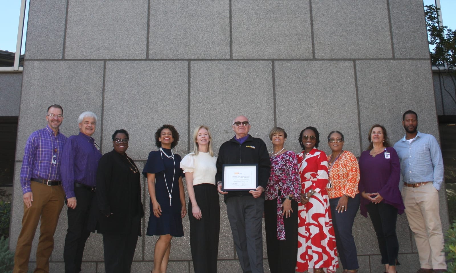 A group of 11 people smile outside of the Louisiana State University Health Sciences Center New Orleans School of Nursing.