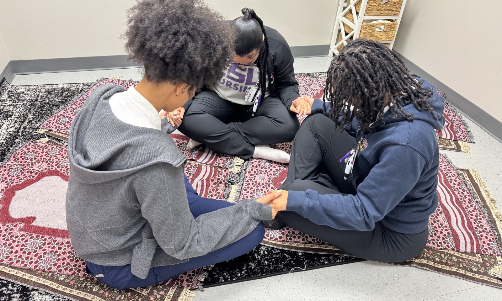 Three LSU Nursing students sit together on patterned prayer mats in the Prayer and Meditation Room, holding hands in a circle during a moment of reflection.