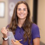 A nursing student wearing a purple LSU School of Nursing shirt smiles while holding a pencil and clipboard in a hallway.