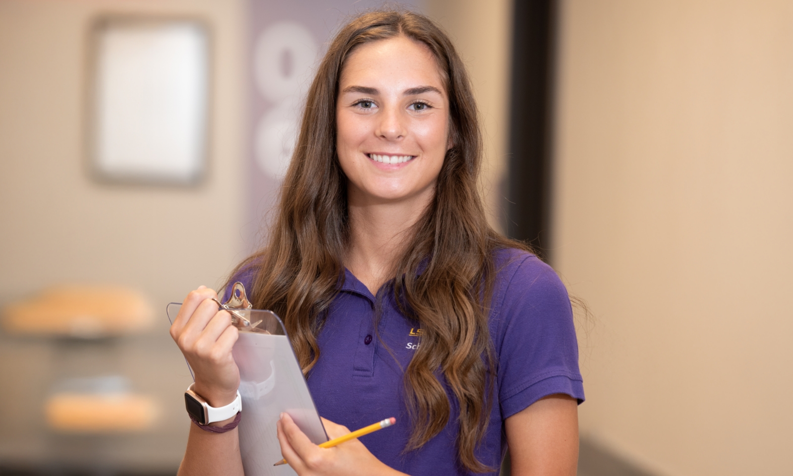 A nursing student wearing a purple LSU School of Nursing shirt smiles while holding a pencil and clipboard in a hallway.
